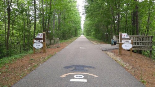 A paved path through a green forest, with signs indicating a turn and a bench on the side.