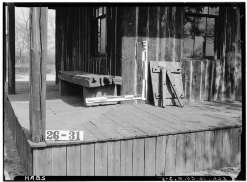 A wooden porch with a bench, two wooden planks, and a number sign, set against a rustic building.