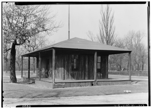 A small, wooden, one-story house with a porch, surrounded by bare trees and grass in a park-like setting.