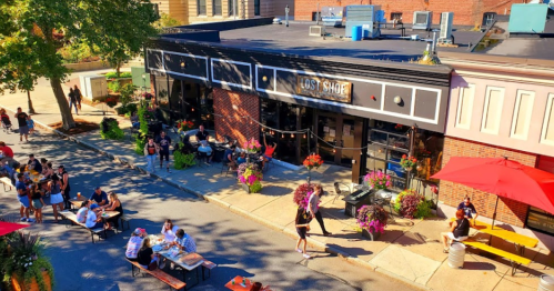 A bustling street scene with outdoor dining, colorful flowers, and people enjoying a sunny day outside a restaurant.