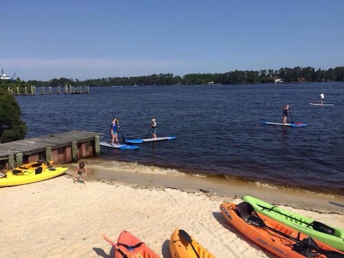 People paddleboarding on a calm lake with kayaks on the sandy shore and trees in the background under a clear blue sky.