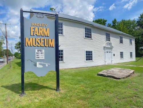 Sign for Hadley Farm Museum in front of a white building, with green grass and a cloudy sky in the background.