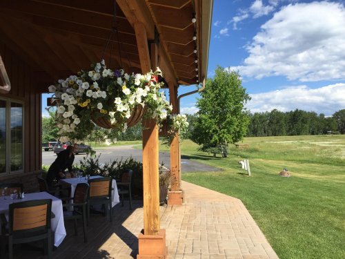 A sunny outdoor dining area with hanging flower baskets, wooden beams, and a view of a green field and blue sky.