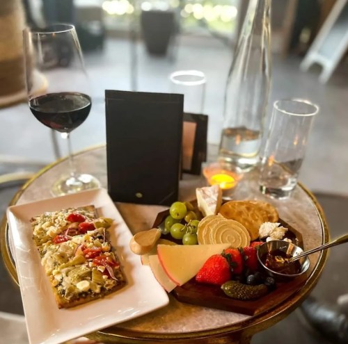 A table setting with a glass of red wine, a plate of gourmet snacks, cheese, fruits, and a menu on a marble surface.