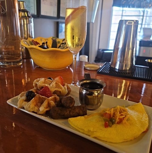 A plate with waffles, sausage, and an omelet, alongside a glass of champagne garnished with a slice of orange.