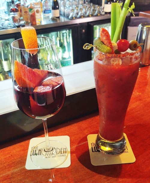 Two colorful cocktails on a bar counter: a red wine spritzer and a Bloody Mary garnished with celery and olives.