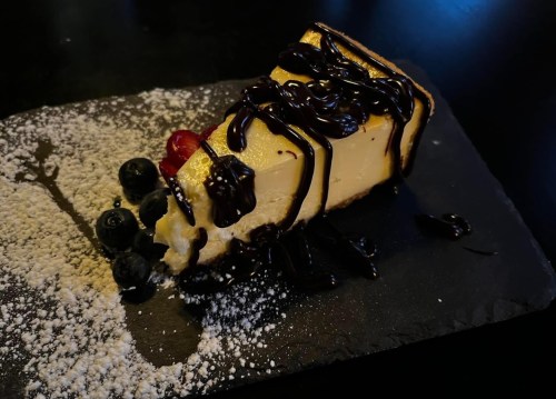 A slice of cheesecake drizzled with chocolate sauce, garnished with berries and powdered sugar on a black slate plate.
