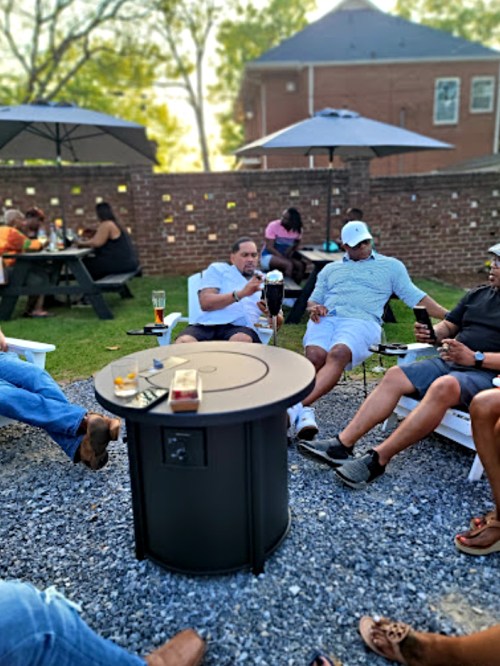 A group of people socializing outdoors, sitting around a fire pit with drinks, under umbrellas in a relaxed setting.