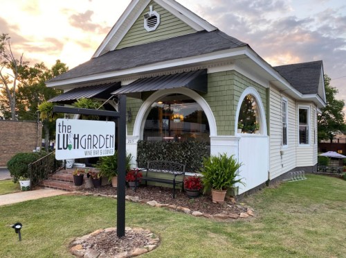 A charming green and white building with a sign reading "the Lush Garden" and a welcoming outdoor seating area.