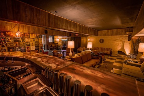 Cozy lounge with wooden walls, retro furniture, and a bar area, featuring warm lighting and a relaxed atmosphere.