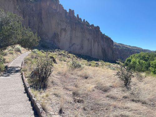 A winding path leads through dry grass and shrubs, with towering cliffs in the background under a clear blue sky.