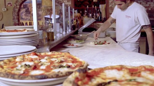 A chef prepares a salad in a pizzeria, with pizzas in the foreground and a rustic kitchen setting.