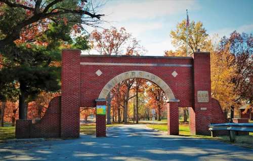 Brick archway entrance to a park, surrounded by colorful autumn trees and a clear blue sky.