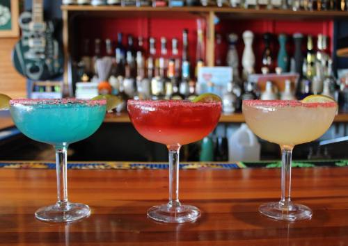 Three colorful margaritas in glassware on a bar, with a variety of drinks and bottles in the background.