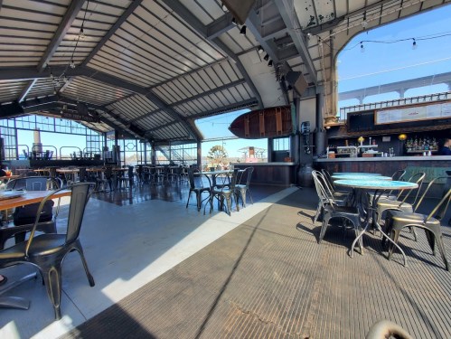 A spacious, modern restaurant interior with metal chairs, large windows, and a bar area, featuring a bright, open atmosphere.