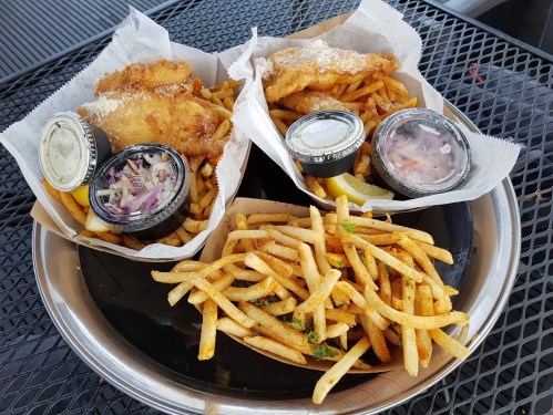 A metal tray with two servings of fish and fries, each with coleslaw and tartar sauce, alongside a pile of extra fries.