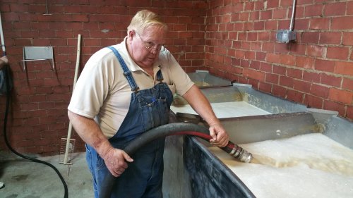 A man in overalls operates a hose, directing liquid into a large tank in a brick-walled facility.