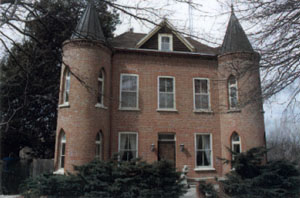 A historic brick house with two conical towers, surrounded by trees and shrubs, on a cloudy day.