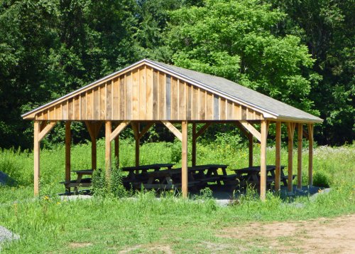 A wooden picnic shelter with a sloped roof, surrounded by green grass and trees in a sunny outdoor setting.