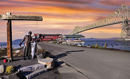 A waterfront scene featuring a statue holding a fish, a gallery sign, and a bridge against a colorful sunset sky.