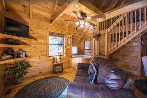 Cozy wooden cabin interior with a sofa, ceiling fan, TV, and a staircase leading to an upper level.