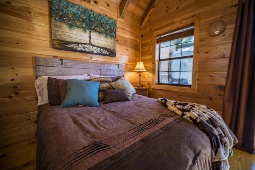 Cozy wooden bedroom with a large bed, decorative pillows, a lamp, and a tree painting on the wall.