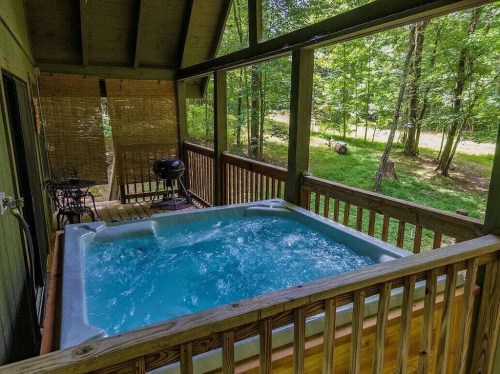 A hot tub on a wooden deck surrounded by trees, with a grill nearby and a serene outdoor view.