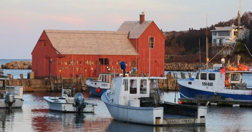 A red barn by the water with fishing boats docked nearby, under a soft sunset sky.