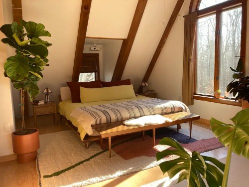 Cozy bedroom with a large bed, wooden beams, plants, and natural light from a window.