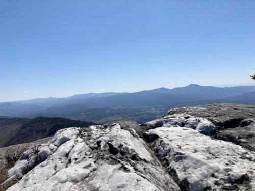 A panoramic view from a rocky summit, showcasing rolling hills and distant mountains under a clear blue sky.