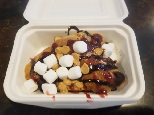 A takeout container filled with dessert featuring pancakes topped with chocolate sauce, marshmallows, and peanut butter chips.