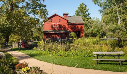 A red barn-style building surrounded by greenery and a pathway, with a picnic table in the foreground.