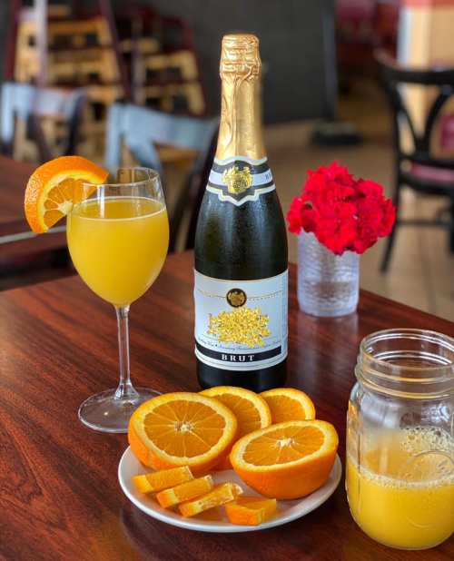 A glass of mimosa with an orange slice, a bottle of sparkling wine, and a plate of sliced oranges on a wooden table.
