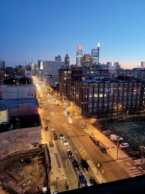 City skyline at dusk, featuring illuminated buildings and streets in Philadelphia, with a clear blue sky above.
