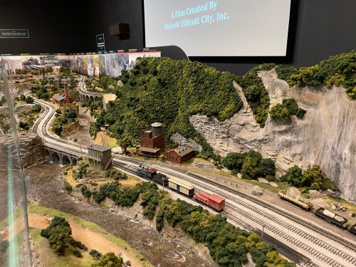 A detailed model train layout featuring tracks, buildings, and lush greenery in a scenic landscape.