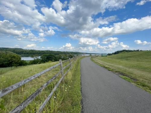A scenic pathway lined with a wooden fence, surrounded by green fields and a river under a partly cloudy sky.