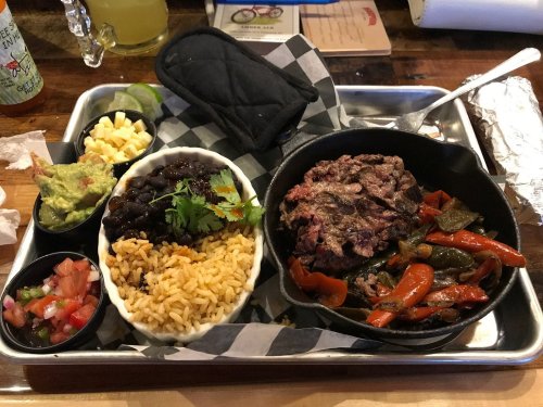 A metal tray with a cast iron skillet of beef, rice, black beans, guacamole, pico de gallo, and grilled peppers.