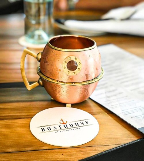 A copper mug sits on a coaster with "Boathouse at the Riverside" printed on it, placed on a wooden table.