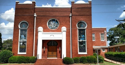 A red brick church with large stained glass windows and a grand entrance, surrounded by greenery and blue skies.