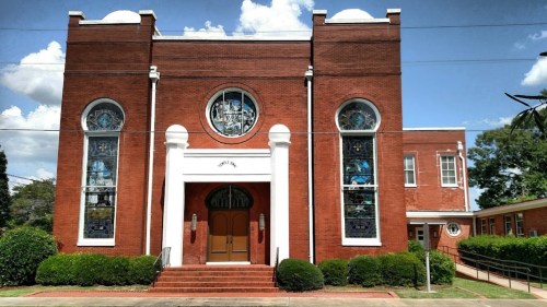 Red brick church with large stained glass windows and a white entrance, surrounded by greenery under a blue sky.