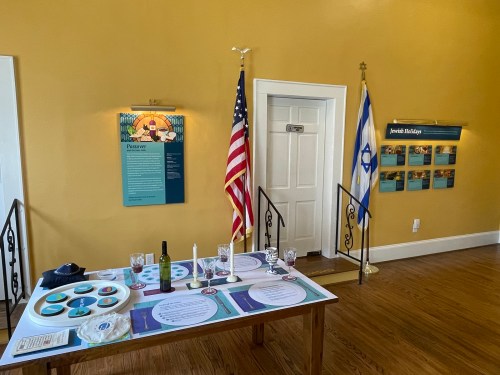 A room with a table set for a Jewish ceremony, featuring flags, a description board, and decorative items.