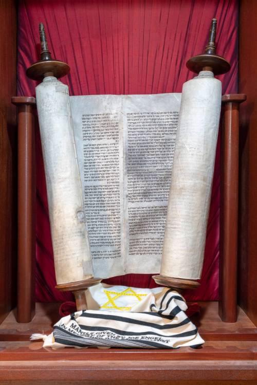 A Torah scroll displayed in a wooden case, with a tallit draped in front, against a red backdrop.