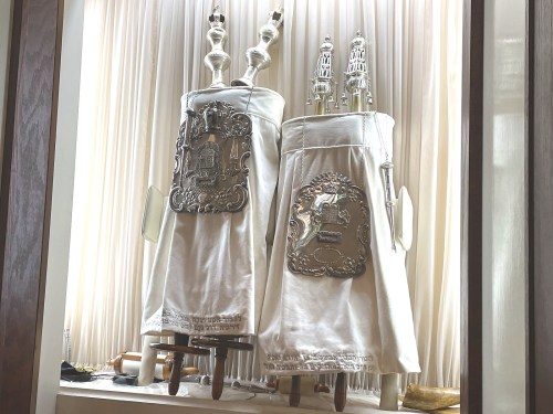 Two ornate Torah scrolls in white covers with silver decorations, displayed against a light backdrop.