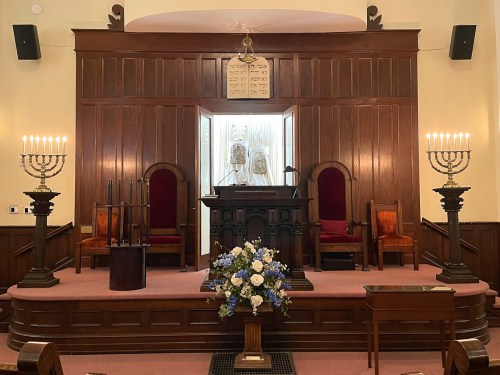 Interior of a synagogue featuring a wooden bimah, two ornate chairs, menorahs, and a floral arrangement.