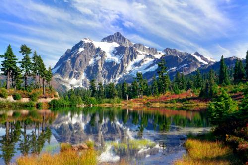 A serene mountain landscape with a snow-capped peak reflecting in a calm lake, surrounded by lush greenery and colorful foliage.