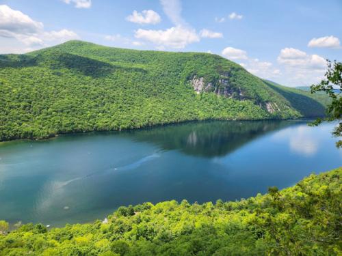 A serene landscape featuring a lush green mountain beside a calm, reflective lake under a blue sky with fluffy clouds.