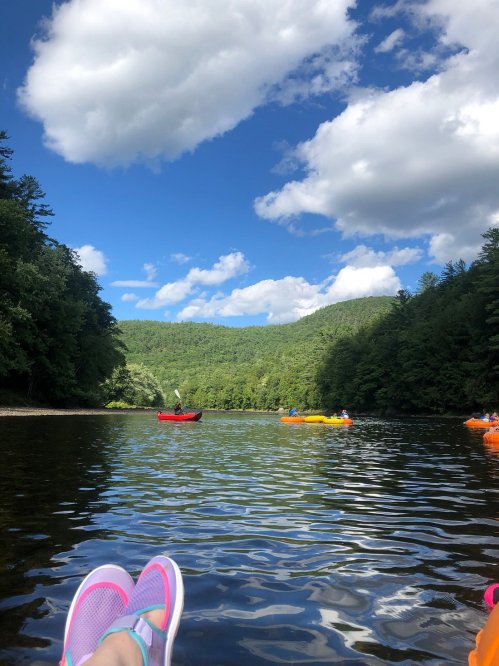 A serene river scene with feet in the foreground, surrounded by lush green hills and kayakers on the water under a blue sky.