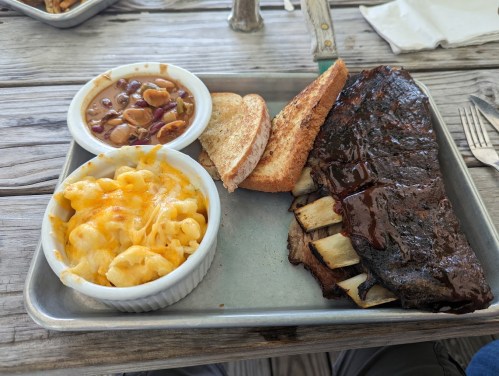 A plate with BBQ ribs, macaroni and cheese, baked beans, and toasted bread on a wooden table.