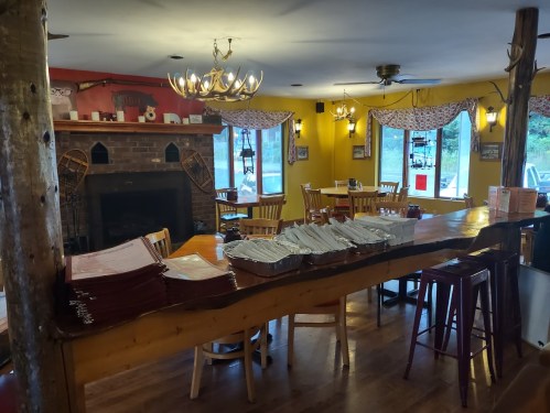 Cozy restaurant interior with wooden tables, a fireplace, and neatly stacked menus on the bar. Bright yellow walls.
