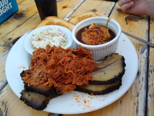 A plate of barbecue featuring pulled pork, brisket slices, coleslaw, and barbecue sauce, with toasted bread on the side.